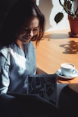 A woman sitting by a wooden table is smiling while looking at a magazine. Sunlight casts a warm glow over the scene, highlighting a cup of coffee with frothy milk on the table. A potted plant adds a touch of green to the background.