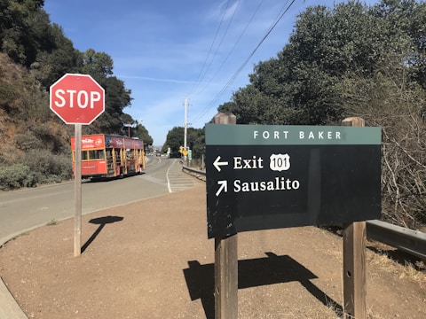 A rural road scene with a stop sign in the foreground and a directional sign indicating routes to Exit 101 and Sausalito. A red tour bus is approaching the stop sign, while lush greenery and trees surround the roadway.