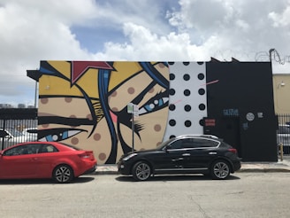 A vibrant mural painted on a building wall features a pop art style with bold lines and bright colors. The composition includes large eyes and colorful patterns, with elements such as polka dots and star shapes. Two cars, one red and one black, are parked along the street in front of the mural. The sky is partly cloudy, adding a contrasting natural background to the urban scene.