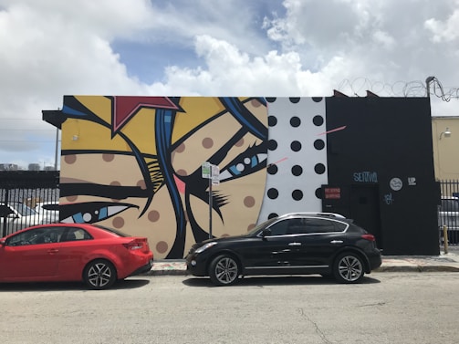 A vibrant mural painted on a building wall features a pop art style with bold lines and bright colors. The composition includes large eyes and colorful patterns, with elements such as polka dots and star shapes. Two cars, one red and one black, are parked along the street in front of the mural. The sky is partly cloudy, adding a contrasting natural background to the urban scene.