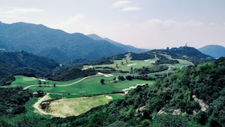 A lush green golf course is nestled among rolling hills and mountains under a partly cloudy sky. The landscape features a combination of manicured lawns, sand traps, and scattered trees, surrounded by dense forests and a mountainous backdrop.