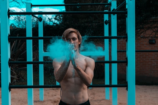 A shirtless person with long hair claps their hands, releasing a cloud of blue powder. They stand in front of a fitness structure with horizontal bars, set outdoors with a background of trees and a brick wall.