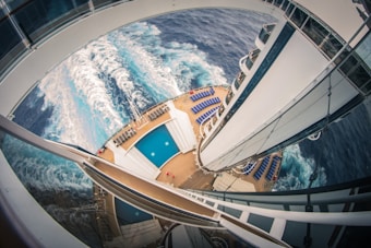 An aerial view of a cruise ship deck featuring two swimming pools surrounded by numerous blue lounge chairs. The ship is sailing through the ocean, creating white waves in the deep blue water. The perspective provides a dynamic angle, emphasizing the curvature of the ship's structures.