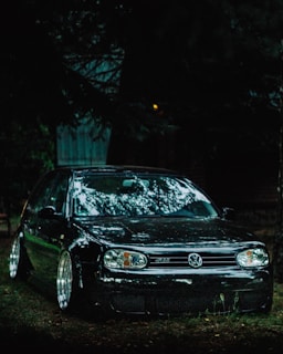 A black Volkswagen car is parked in a dimly lit area surrounded by trees, with reflections visible on its glossy surface. The vehicle has modified wheels and a sleek design.