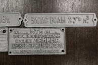 The image contains three vintage metal plaques with Russian inscriptions. The plaques appear to be historical or industrial, possibly from equipment or machinery. They are laid out against a wooden background.