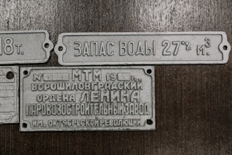 The image contains three vintage metal plaques with Russian inscriptions. The plaques appear to be historical or industrial, possibly from equipment or machinery. They are laid out against a wooden background.