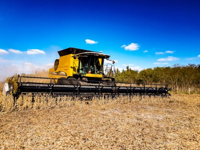 A large, yellow agricultural combine harvester operates in a field, cutting and collecting crops. The machine's wide header attachment dominates the foreground, while the background features a clear blue sky with sparse clouds and a line of trees.