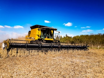 A large, yellow agricultural combine harvester operates in a field, cutting and collecting crops. The machine's wide header attachment dominates the foreground, while the background features a clear blue sky with sparse clouds and a line of trees.