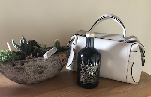 A white handbag is placed on a wooden surface next to a decorative concrete planter filled with a variety of succulents and small cacti. In front of the handbag is a bottle labeled 'Hustle', featuring an intricate gold and black design. The plain background enhances the focus on these objects.