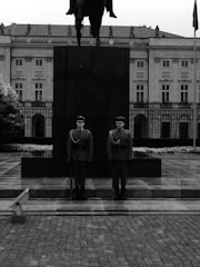 Two uniformed guards stand on a paved area in front of a large, dark monument with an equestrian statue atop it. The backdrop is a grand, classical building with ornate architectural details, including columns and rows of windows.