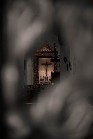 The image captures a view of a church interior with an ornate altar in the background. Candles are lit on the altar, and a crucifix is prominently displayed. The scene is partially obscured, framed by blurred elements in the foreground, creating a sense of peeking into the sacred space.