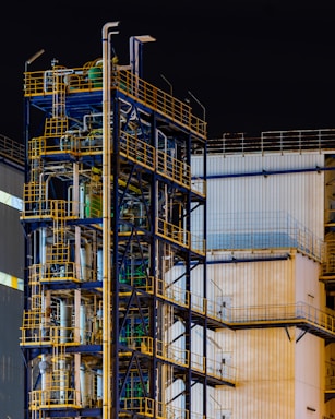 A complex industrial structure featuring multiple levels of metal platforms, pipes, and railings against the backdrop of a corrugated building. The structure is primarily composed of blue and yellow elements with various industrial components visible.
