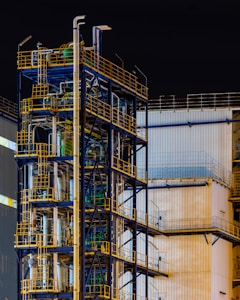A complex industrial structure featuring multiple levels of metal platforms, pipes, and railings against the backdrop of a corrugated building. The structure is primarily composed of blue and yellow elements with various industrial components visible.