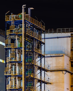 A complex industrial structure featuring multiple levels of metal platforms, pipes, and railings against the backdrop of a corrugated building. The structure is primarily composed of blue and yellow elements with various industrial components visible.