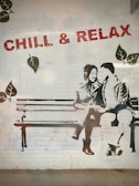 A stencil-style mural on a brick wall depicts two people sitting closely on a bench, sharing a lighthearted moment. The phrase 'CHILL & RELAX' is stenciled above in bold red letters, and leaves are painted around the scene, adding a nature-themed element.