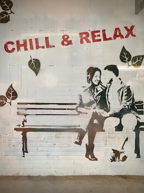 A stencil-style mural on a brick wall depicts two people sitting closely on a bench, sharing a lighthearted moment. The phrase 'CHILL & RELAX' is stenciled above in bold red letters, and leaves are painted around the scene, adding a nature-themed element.