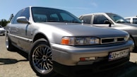 A silver Honda sedan with a classic design is parked outdoors, with other cars visible in the background. The car's body is shiny and the wheels have detailed rims.