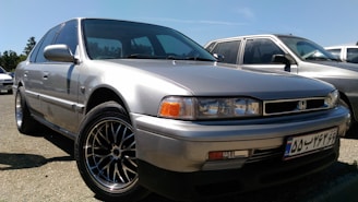 A silver Honda sedan with a classic design is parked outdoors, with other cars visible in the background. The car's body is shiny and the wheels have detailed rims.