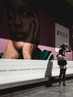 A large advertisement displays a close-up of a person wearing glasses, with a focus on their contemplative expression. In front of this poster, a woman stands in sunlight, casting a shadow on the ground. She appears to be adjusting her hair, carrying a bag on her shoulder, and wearing a short dress.