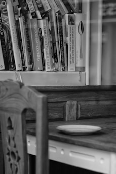 A monochromatic image of a wooden shelf filled with various cookbooks. Nearby, a wooden chair with intricate carvings sits next to a wooden table. A simple, empty plate rests on the tabletop, adding to the stillness of the scene.