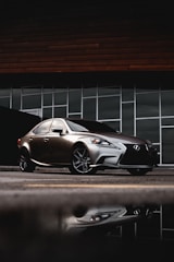A sleek, silver luxury sedan is parked in front of a modern building with large rectangular glass windows. The car's reflection is visible on the wet pavement, and the backdrop includes a dark, wood-paneled facade.