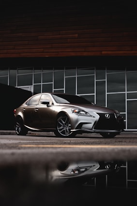 A sleek, silver luxury sedan is parked in front of a modern building with large rectangular glass windows. The car's reflection is visible on the wet pavement, and the backdrop includes a dark, wood-paneled facade.