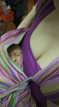 A sleeping baby is nestled closely against an adult who is likely its parent, wrapped in a colorful, striped fabric. The dominant fabric colors include pink, purple, and green, providing a cozy and secure environment for the baby.