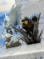Two detailed figurines from the Monster Hunter Iceborne series are displayed prominently on a reflective surface. One figure resembles a majestic, icy dragon with expansive wings, while the other is a dark, rugged creature perched on rocky terrain. The background features the Monster Hunter Iceborne logo and name.