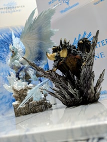 Two detailed figurines from the Monster Hunter Iceborne series are displayed prominently on a reflective surface. One figure resembles a majestic, icy dragon with expansive wings, while the other is a dark, rugged creature perched on rocky terrain. The background features the Monster Hunter Iceborne logo and name.