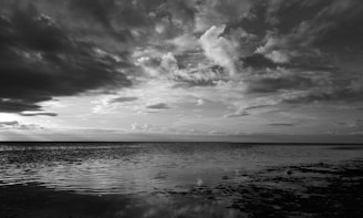 A tranquil seascape features a vast expanse of water beneath a dramatic sky filled with thick, textured clouds. The water appears calm, with gentle ripples reflecting the gradient of the sky above. Horizon stretches seamlessly, meeting the sky in a distant line.