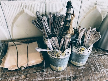 A rustic setting with two metal containers holding ornate silverware on a wooden surface. Beside them, a stack of brown paper napkins is neatly arranged within a wire basket. The containers have a vintage, distressed look with a lace-like design around the top.