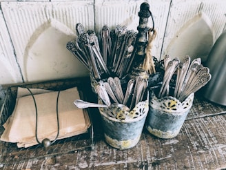 A rustic setting with two metal containers holding ornate silverware on a wooden surface. Beside them, a stack of brown paper napkins is neatly arranged within a wire basket. The containers have a vintage, distressed look with a lace-like design around the top.