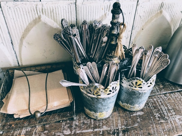 A rustic setting with two metal containers holding ornate silverware on a wooden surface. Beside them, a stack of brown paper napkins is neatly arranged within a wire basket. The containers have a vintage, distressed look with a lace-like design around the top.