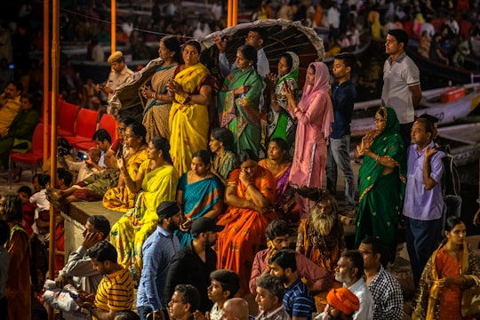 A group of people gathered in colorful traditional attire, engaged in a ritual or prayer at a public event or ceremony. The crowd is diverse, with a mix of men and women, some sitting while others stand with folded hands. The atmosphere suggests a cultural or religious significance.