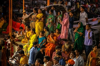 A group of people gathered in colorful traditional attire, engaged in a ritual or prayer at a public event or ceremony. The crowd is diverse, with a mix of men and women, some sitting while others stand with folded hands. The atmosphere suggests a cultural or religious significance.