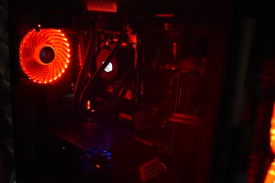 A close-up view of the interior of a computer case with illuminated components. The most prominent feature is a red-lit cooling fan on the left side, emitting a bright glow. Various components including cables and a central processing unit cooler are visible, shrouded in shadows.