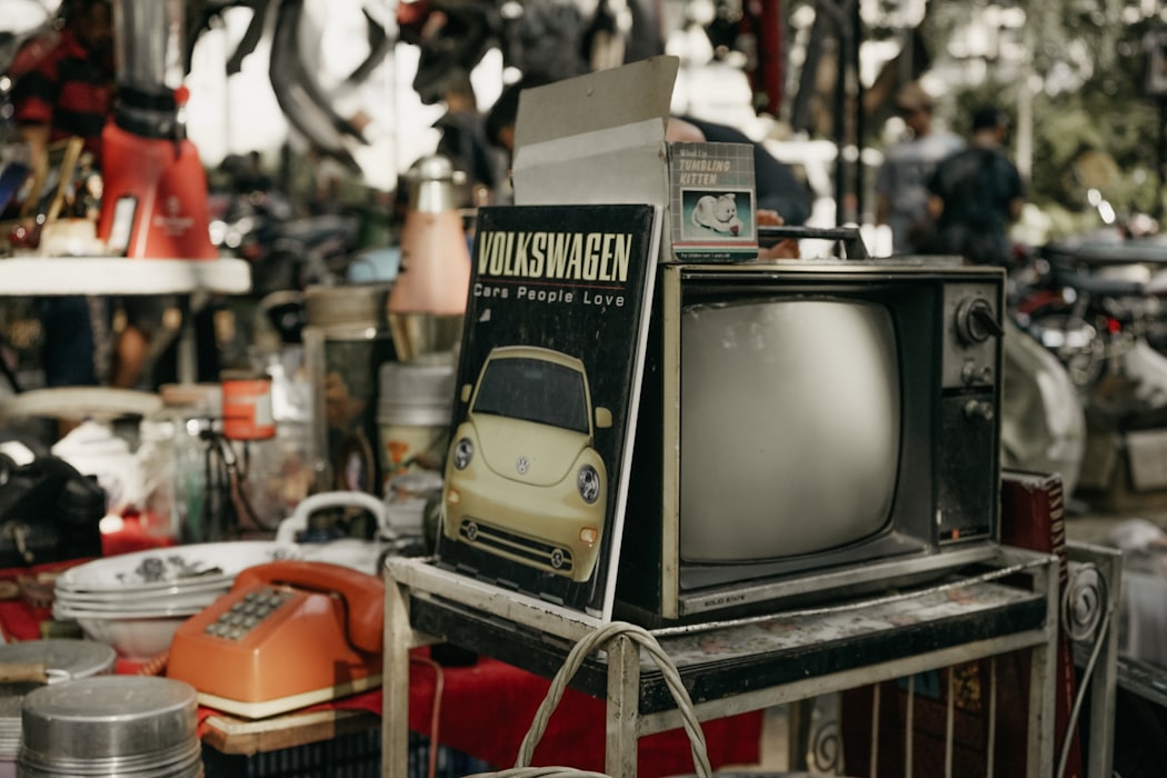 Thrift store with various items on the table including an old television in the foreground, with an old Volkswagen book leaned up against it. 