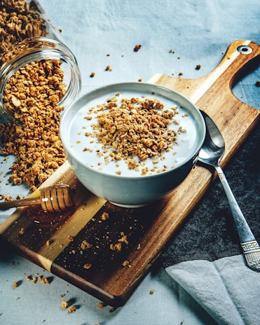 A bowl filled with yogurt topped with granola sits on a wooden cutting board. Granola is scattered around the board, and a jar lays on its side with more granola spilling out. A honey dipper rests near the bowl. The setting includes a spoon and a dark napkin, creating a cozy breakfast scene.