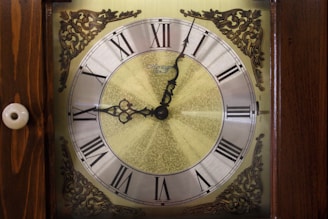 An ornate clock face with Roman numerals is surrounded by intricate brass designs at the corners. The clock hands are decorative and point to just past four o'clock. The background has a gradient texture with a metallic gold hue.