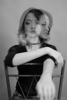 A monochrome photograph of a woman sitting on a chair showing a double exposure effect. Her face is duplicated to the side, creating a blurred and surreal visual impact. She has shoulder-length hair, and her arms are resting on the back of the chair with an outstretched gesture.