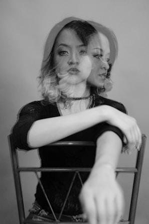 A monochrome photograph of a woman sitting on a chair showing a double exposure effect. Her face is duplicated to the side, creating a blurred and surreal visual impact. She has shoulder-length hair, and her arms are resting on the back of the chair with an outstretched gesture.