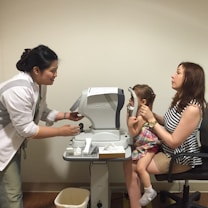 A woman in a white coat is assisting a young child sitting on another woman's lap during an eye examination. The child is positioned in front of a medical device for eye testing, with the woman holding her in place. The setting appears to be an eye clinic room.