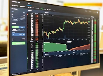 A computer monitor displays a cryptocurrency exchange interface with various graphs, charts, and numerical data. On the left side, there are options for buying and selling, as well as a list of recent trades. The main graph on the right shows a price chart with a candlestick pattern, indicating fluctuations in the market over a period of time.