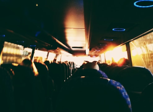 The interior of a bus with several passengers seated in rows. Warm sunlight streams through the windows, creating a cozy atmosphere. The ceiling features circular blue LED lights.