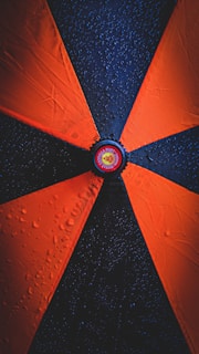 A close-up view of an umbrella with alternating orange and black panels, each segment adorned with water droplets. The center of the umbrella features a circular logo with text and an emblem.