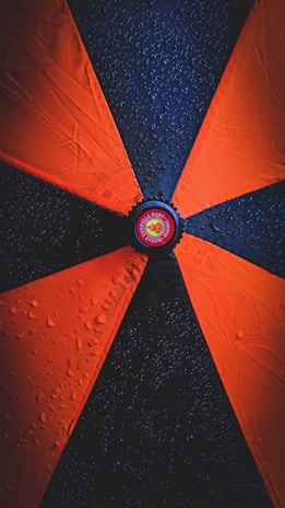 A close-up view of an umbrella with alternating orange and black panels, each segment adorned with water droplets. The center of the umbrella features a circular logo with text and an emblem.