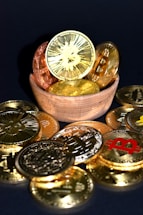 A wooden bowl filled with various golden and copper-colored cryptocurrency coins is surrounded by more coins on a dark surface. Each coin features distinct cryptocurrency symbols, including Bitcoin, Ethereum, and others.