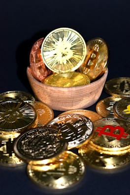 A wooden bowl filled with various golden and copper-colored cryptocurrency coins is surrounded by more coins on a dark surface. Each coin features distinct cryptocurrency symbols, including Bitcoin, Ethereum, and others.