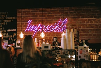 A cozy coffee shop scene with exposed brick walls and warm lighting. A neon sign reading 'Impossible' is prominently displayed above a shelf stocked with various cups and products. Several people are standing near the counter, and there is a menu board on the wall.