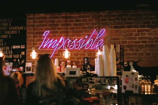 A cozy coffee shop scene with exposed brick walls and warm lighting. A neon sign reading 'Impossible' is prominently displayed above a shelf stocked with various cups and products. Several people are standing near the counter, and there is a menu board on the wall.